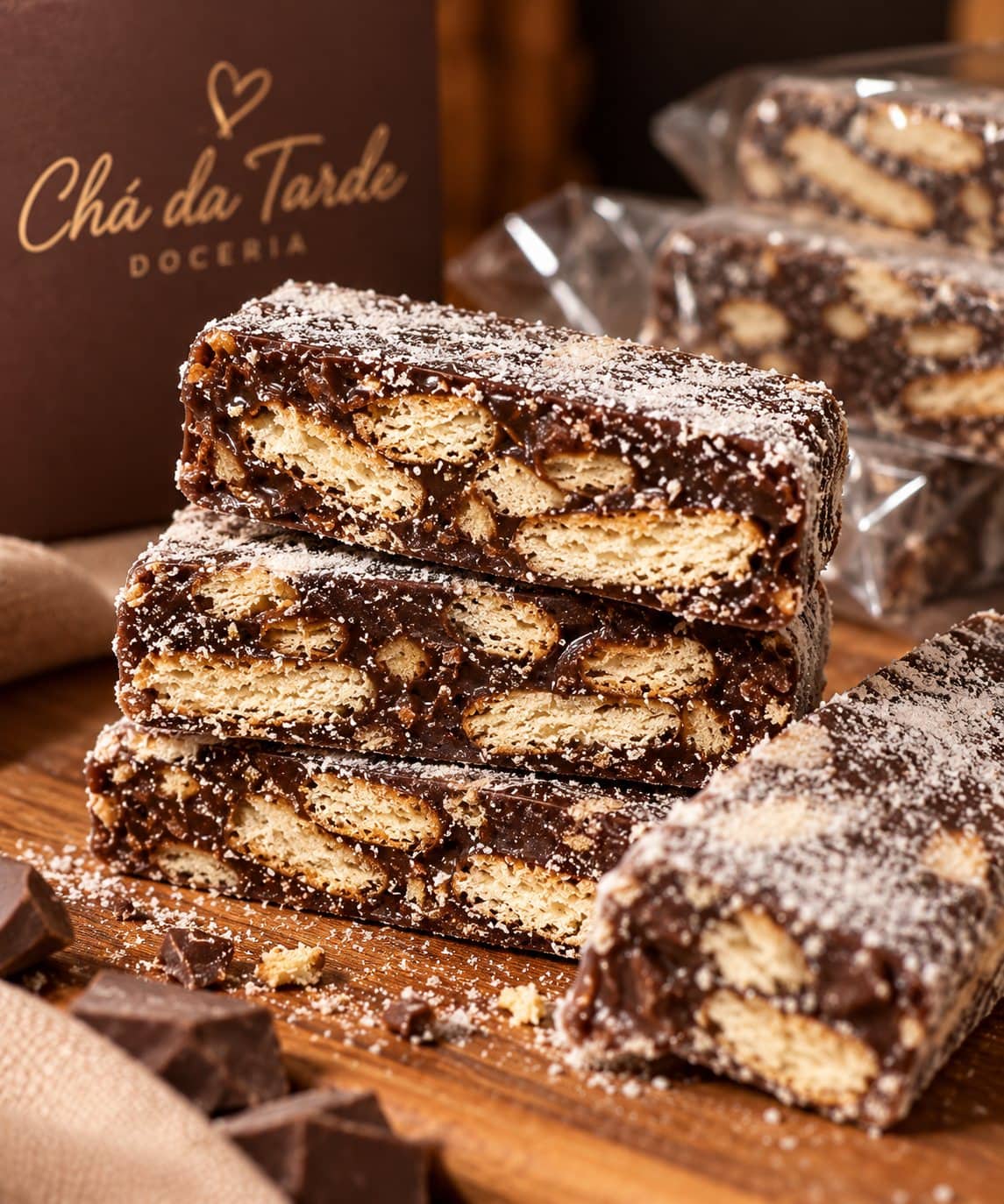 Palhas italianas empilhadas, com pedaços de biscoito visíveis no corte do chocolate, sobre madeira e ao lado da caixa da Chá da Tarde